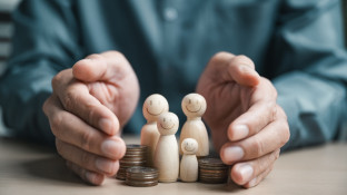 Een paar handen beschermt een houten poppetjesgezin en stapels munten op een tafel
